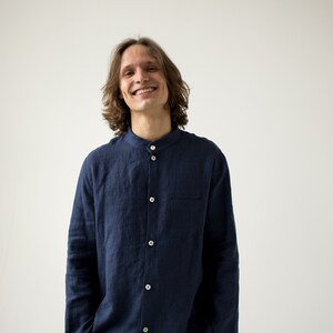May include: A navy blue linen shirt with a mandarin collar and matching pants. The shirt has white buttons and a chest pocket. The person is smiling. The outfit is casual and comfortable.