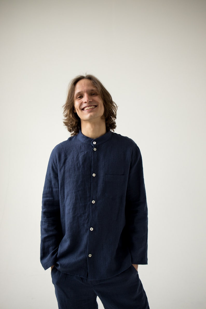 May include: A navy blue linen shirt with a mandarin collar and matching pants. The shirt has white buttons and a chest pocket. The person is smiling. The outfit is casual and comfortable.