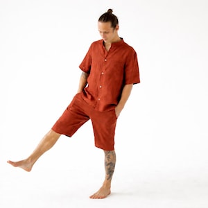 May include: A rust-colored linen shirt and shorts set. The short-sleeved shirt has a mandarin collar and button-down front. The shorts are knee-length. The outfit is worn by a person with a topknot hairstyle.