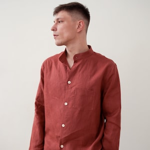 May include: A rust-colored linen shirt with a mandarin collar and long sleeves. The shirt has a single chest pocket and is buttoned up. The matching pants are visible at the bottom of the image. The outfit is made of a lightweight fabric.