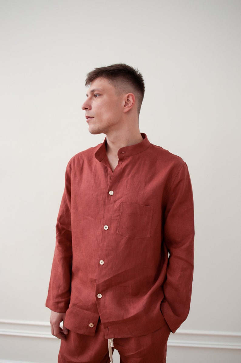 May include: A rust-colored linen shirt with a mandarin collar and long sleeves. The shirt has a single chest pocket and is buttoned up. The matching pants are visible at the bottom of the image. The outfit is made of a lightweight fabric.