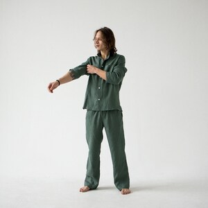 May include: A green linen pajama set. The set includes a button-down shirt with a collar and long pants. The person in the image is rolling up the sleeves of the shirt. The background is plain white.