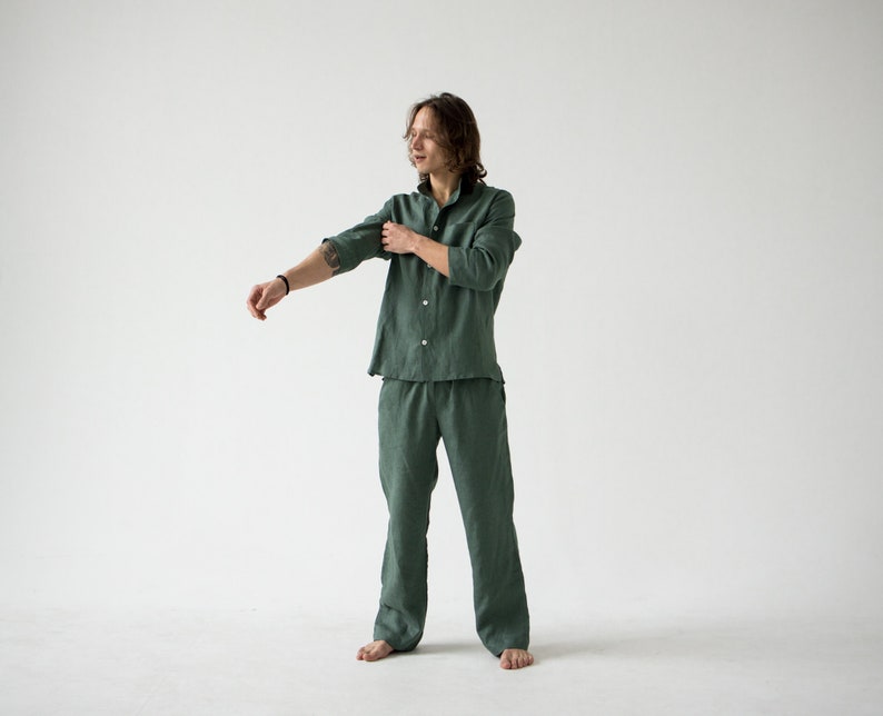 May include: A green linen pajama set. The set includes a button-down shirt with a collar and long pants. The person in the image is rolling up the sleeves of the shirt. The background is plain white.