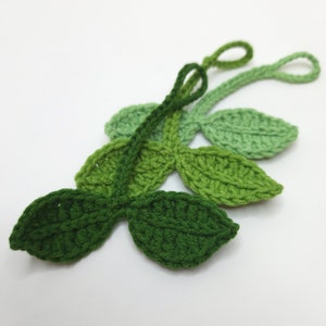 May include: Crocheted leaf embellishments in varying shades of green. The leaves are arranged in a cluster, with a loop at the end of each stem. The handmade leaves are suitable for crafts and decorations.