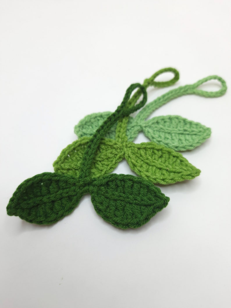 May include: Crocheted leaf embellishments in varying shades of green. The leaves are arranged in a cluster, with a loop at the end of each stem. The handmade leaves are suitable for crafts and decorations.