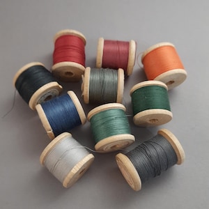 May include: A selection of colourful thread spools. The wooden spools are filled with thread in shades of red, orange, green, blue, and grey. The spools are arranged on a grey surface.