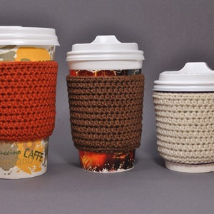 May include: Three coffee cups with crocheted cup cozies in different colors. The cozies are orange, brown, and beige.