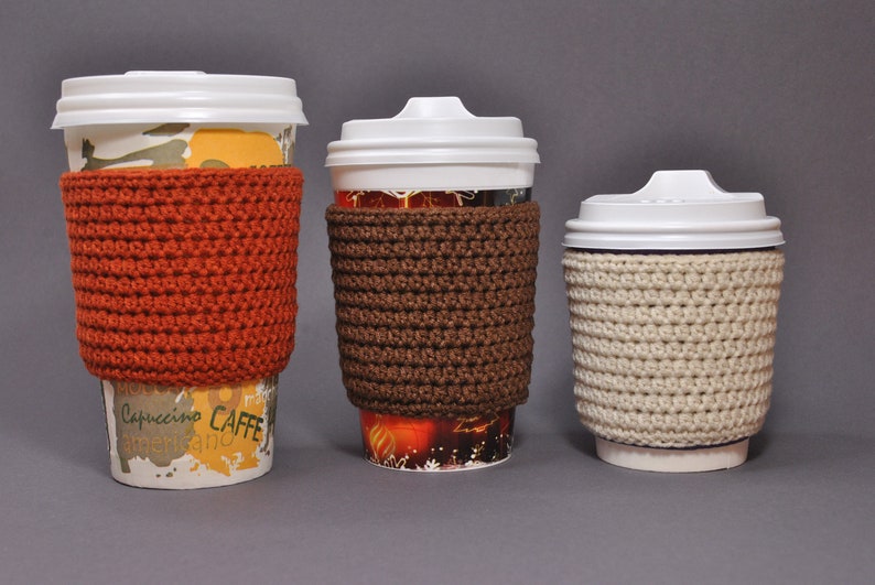 May include: Three coffee cups with crocheted cup cozies in different colors. The cozies are orange, brown, and beige.