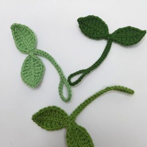 May include: Three crocheted leaf sprigs in varying shades of green. Each sprig features two leaves and a stem, with the leaves displaying a textured pattern. The leaves are in shades of light, medium, and dark green.