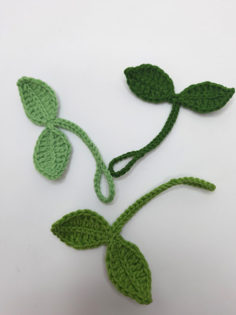 May include: Three crocheted leaf sprigs in varying shades of green. Each sprig features two leaves and a stem, with the leaves displaying a textured pattern. The leaves are in shades of light, medium, and dark green.