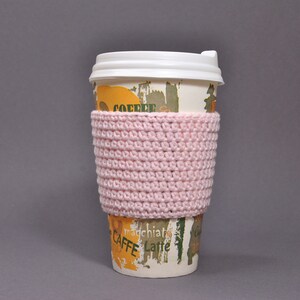 May include: A pink crocheted coffee cup cozy. The cozy is on a disposable coffee cup with the words "CAFFE" and "latte" printed on it.