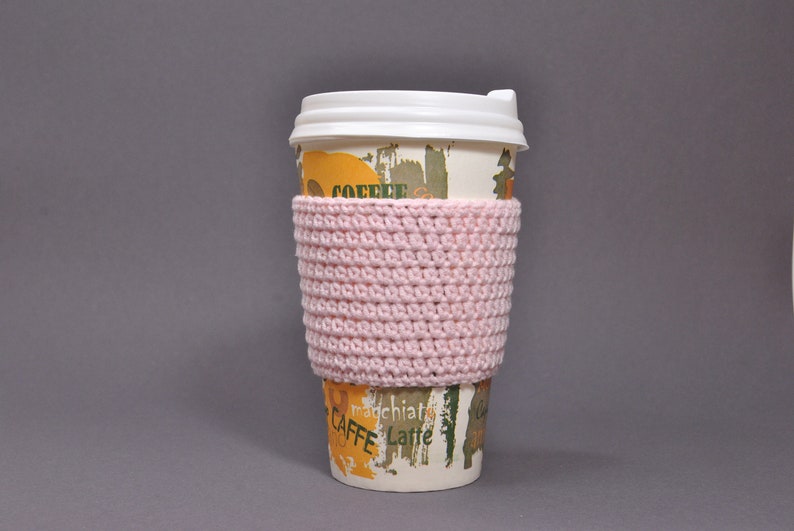 May include: A pink crocheted coffee cup cozy. The cozy is on a disposable coffee cup with the words "CAFFE" and "latte" printed on it.