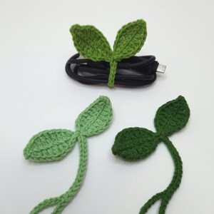 May include: Crocheted leaf cable organizers in shades of green. One organizer is wrapped around a black cable. Two additional leaf organizers are displayed below. These handmade accessories are designed to keep cables tidy and add a touch of nature.