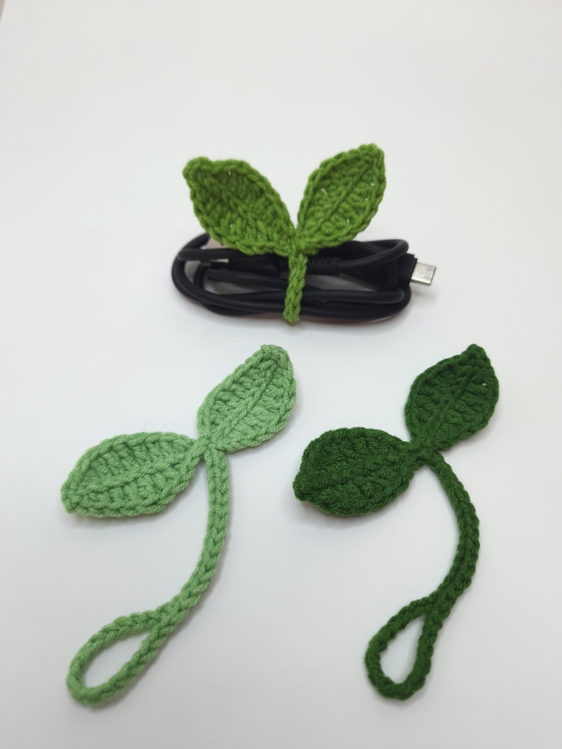 May include: Crocheted leaf cable organizers in shades of green. One organizer is wrapped around a black cable. Two additional leaf organizers are displayed below. These handmade accessories are designed to keep cables tidy and add a touch of nature.