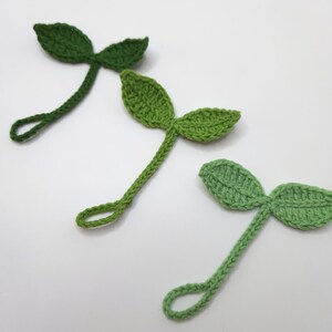 May include: Three handmade crochet leaf embellishments in varying shades of green. Each leaf has two rounded lobes and a long stem with a loop at the end. The leaves are arranged diagonally against a white background.