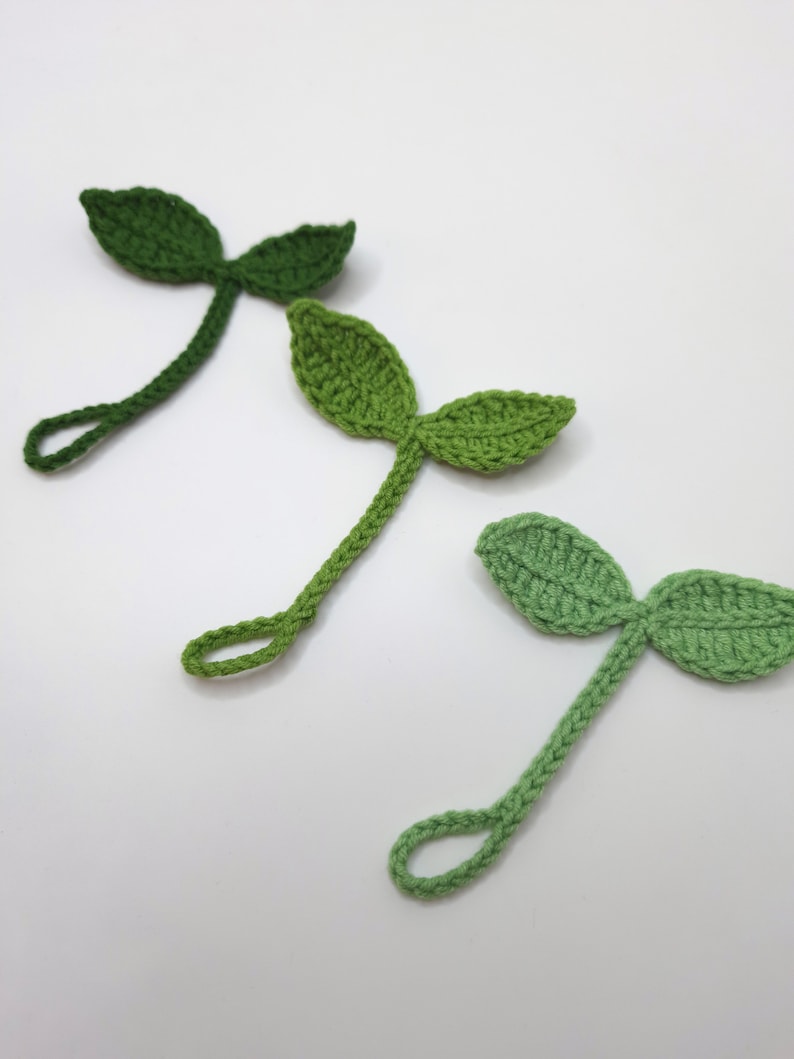 May include: Three handmade crochet leaf embellishments in varying shades of green. Each leaf has two rounded lobes and a long stem with a loop at the end. The leaves are arranged diagonally against a white background.