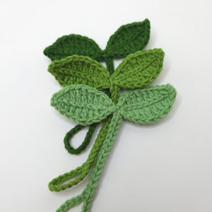 May include: Crocheted green leaf embellishments in varying shades of green. The leaves are arranged in a layered pattern, with each leaf featuring a detailed, textured stitch. The stems have loops at the end. These handmade leaves are perfect for craft projects.