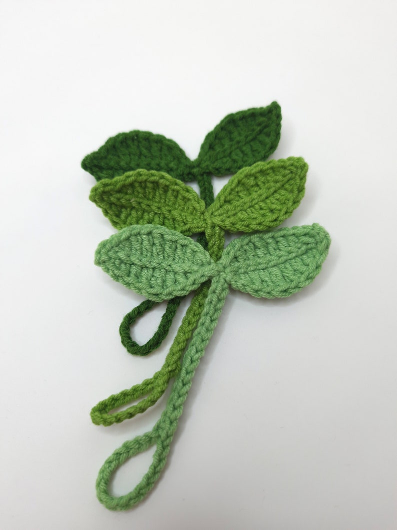 May include: Crocheted green leaf embellishments in varying shades of green. The leaves are arranged in a layered pattern, with each leaf featuring a detailed, textured stitch. The stems have loops at the end. These handmade leaves are perfect for craft projects.