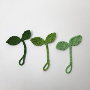 May include: Three crocheted green sprout shapes with two leaves each. The sprouts are in varying shades of green, from dark to light. Each sprout has a looped stem, and they are arranged against a white background.