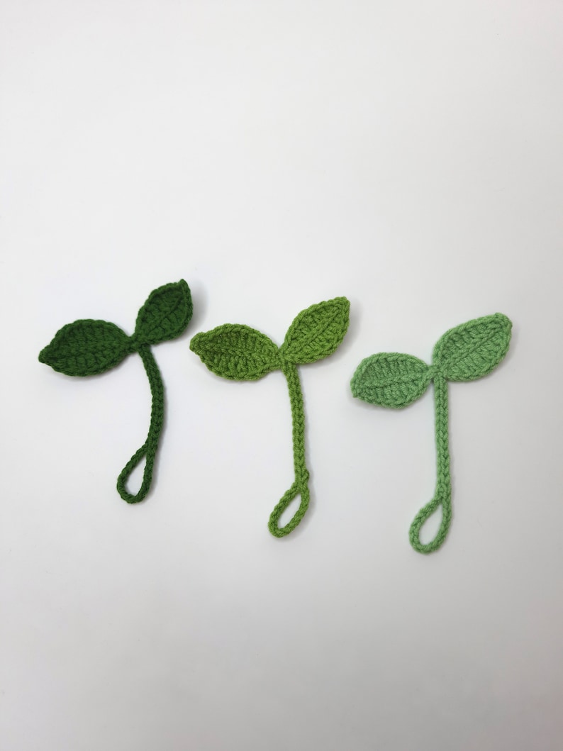 May include: Three crocheted green sprout shapes with two leaves each. The sprouts are in varying shades of green, from dark to light. Each sprout has a looped stem, and they are arranged against a white background.