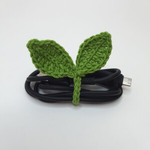 May include: A black USB cable is neatly bundled with a handmade, green crochet leaf tie. The leaf tie features two leaves and a stem, adding a touch of nature to the tech accessory. The background is plain white.