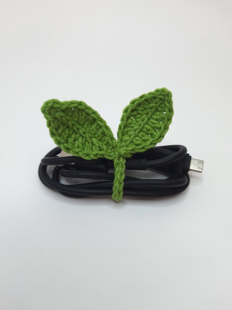 May include: A black USB cable is neatly bundled with a handmade, green crochet leaf tie. The leaf tie features two leaves and a stem, adding a touch of nature to the tech accessory. The background is plain white.