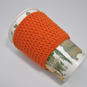 May include: Orange crocheted coffee cup cozy on a white paper cup with the words "Mocca, Cappuccino, and Latte" printed on it.