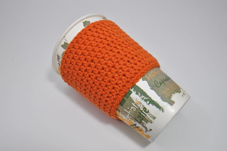May include: Orange crocheted coffee cup cozy on a white paper cup with the words "Mocca, Cappuccino, and Latte" printed on it.