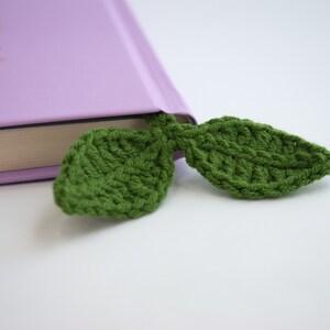 May include: A green, crocheted leaf bookmark is inserted into a purple book. The bookmark is made of a textured yarn, and the leaves are detailed with visible stitching. The book's pages are visible, and the background is white.