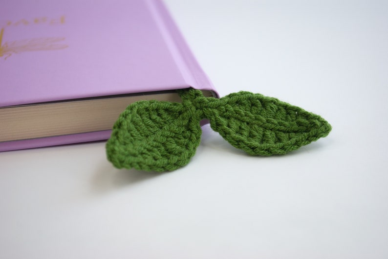 May include: A green, crocheted leaf bookmark is inserted into a purple book. The bookmark is made of a textured yarn, and the leaves are detailed with visible stitching. The book's pages are visible, and the background is white.