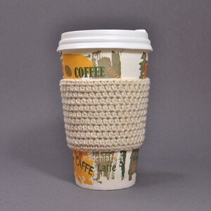 May include: A white coffee cup with a crocheted beige cup cozy. The cup has a colorful design with the word "Coffee" and "Caffe" printed on it.