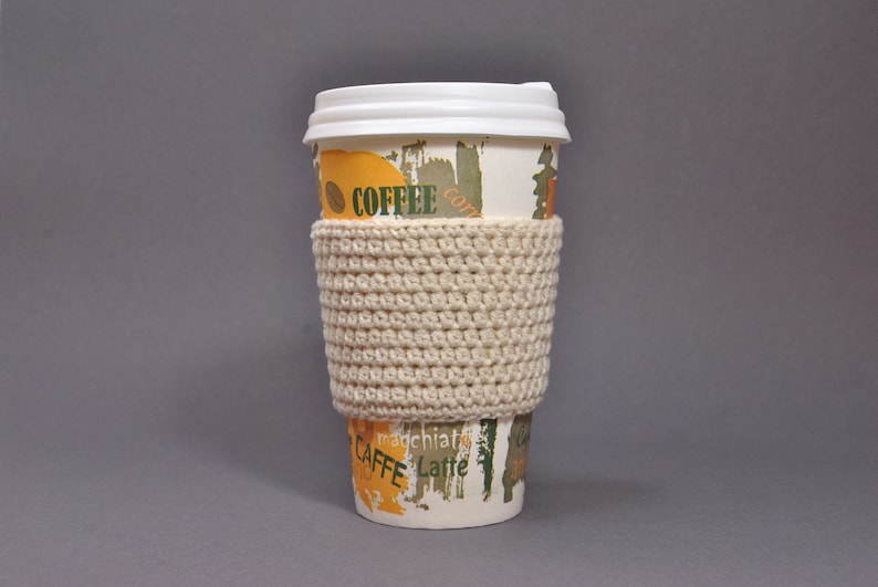 May include: A white coffee cup with a crocheted beige cup cozy. The cup has a colorful design with the word "Coffee" and "Caffe" printed on it.