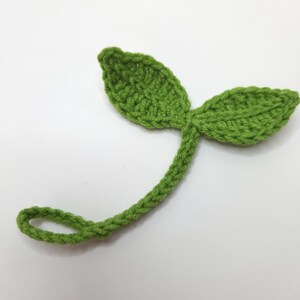 May include: A handmade, green crochet leaf with two leaves and a stem. The stem curves into a loop. The crochet work is visible, showing the texture of the yarn. The leaf is a bright, spring green color.