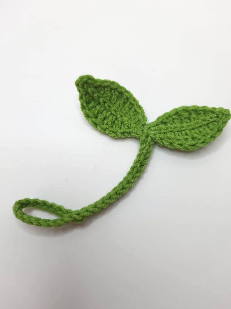 May include: A handmade, green crochet leaf with two leaves and a stem. The stem curves into a loop. The crochet work is visible, showing the texture of the yarn. The leaf is a bright, spring green color.