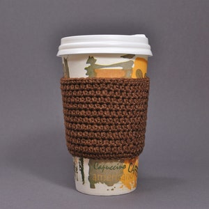 May include: A brown crocheted coffee cup cozy with a white paper cup with a yellow and green design. The cup has the text "Cappuccino Cafe" on it.