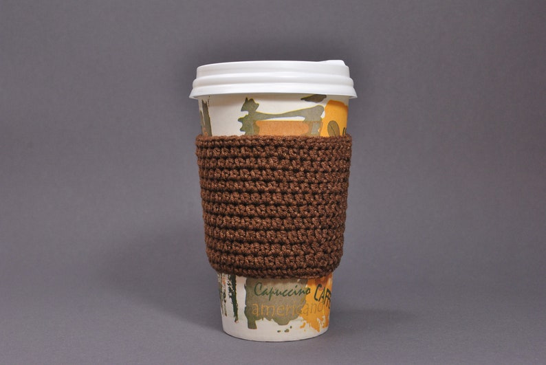 May include: A brown crocheted coffee cup cozy with a white paper cup with a yellow and green design. The cup has the text "Cappuccino Cafe" on it.