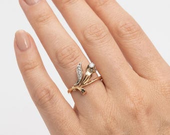May include: A gold ring featuring a floral-inspired design with a single round brilliant-cut diamond set at the top and a curved leaf-shaped accent adorned with a row of small pavé-set diamonds, all crafted in a polished yellow gold band.