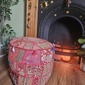 May include: A round, red patchwork pouf with detailed embroidery and tassels. It sits on a wooden floor, in front of a fireplace with a black mantel and gold star decorations. The style is bohemian, with a global influence.