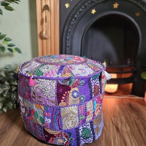 May include: A square, patchwork pouf in shades of purple, pink, and gold. The pouf is covered in various fabric patterns and textures, with tassels on the sides. The decorative piece is placed on a wooden floor.