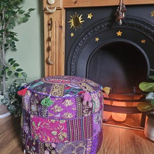 May include: A round, patchwork pouf in shades of purple, pink, and gold. The pouf is adorned with tassels and intricate embroidered patterns. It sits on a wooden floor, with a fireplace and decorative elements in the background.