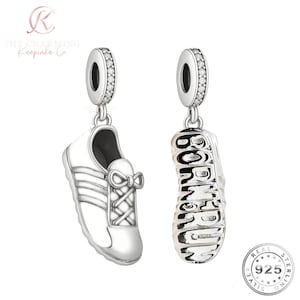 May include: Two silver charms shaped like sneakers. One charm is a side view of a sneaker with laces and a bow. The other charm is the sole of a sneaker with the word "BORN TO RUN" engraved. Both charms have a sparkling top.