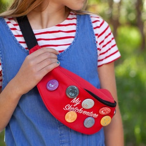 May include: A red fanny pack with a white "My Sketchventure" inscription and colorful cartoon character pins. The fanny pack is worn by a person wearing a blue overall dress and a red and white striped shirt.