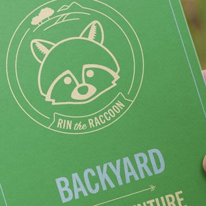 Peut inclure: Un livre vert avec une illustration de raton laveur et le texte "Rin the Raccoon Backyard Adventure" sur la couverture.