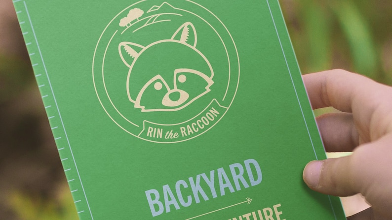 Peut inclure: Un livre vert avec une illustration de raton laveur et le texte "Rin the Raccoon Backyard Adventure" sur la couverture.