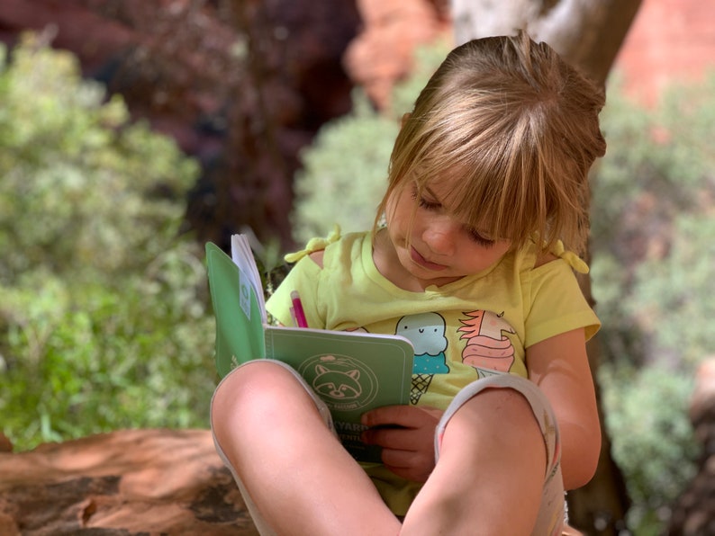 Peut inclure: Une jeune personne est assise &agrave; l'ext&eacute;rieur, en train d'&eacute;crire dans un journal vert avec un crayon rose. Le journal a une illustration de raton laveur sur la couverture et le texte "Nature's Diary".