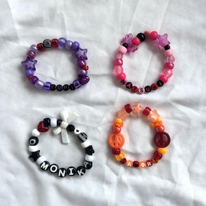 Puede incluir: Cuatro pulseras de cuentas de colores. Una es morada con cuentas de estrella y el nombre "YURI". Otra es rosa con cuentas de estrella y el nombre "NATSUKI". Una tercera es blanca y negra con el nombre "MONIKA". La última es naranja con un signo de la paz y el nombre "SAYORI".