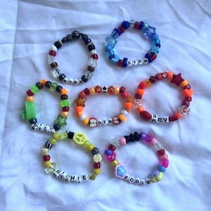 Puede incluir: Una colección de pulseras de cuentas de colores con varios diseños y nombres escritos con cuentas de letras. Las pulseras presentan una mezcla de cuentas redondas, en forma de estrella y de flor en tonos de azul, rojo, naranja, amarillo y verde.