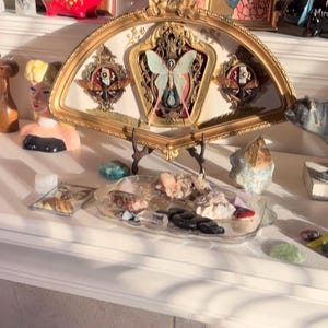 May include: A decorative display featuring a gold-framed fan with a butterfly and religious imagery. The display is on a white surface, surrounded by crystals, figurines, and a red pig-shaped bank. The fan is approximately 46 cm wide.