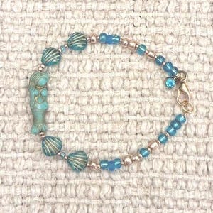Peut inclure: Un bracelet de perles composé d'une breloque sirène turquoise, de plusieurs perles en forme de coquillage et d'une alternance de petites perles bleues et dorées métallisées. Le bracelet comporte un fermoir mousqueton doré et une breloque cristal.