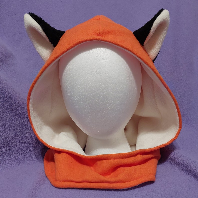 A faceless mannequin wearing a detached orange hood.  The hood has black fox ears with ivory in front.  The same ivory fleece lines the inside of the hood.